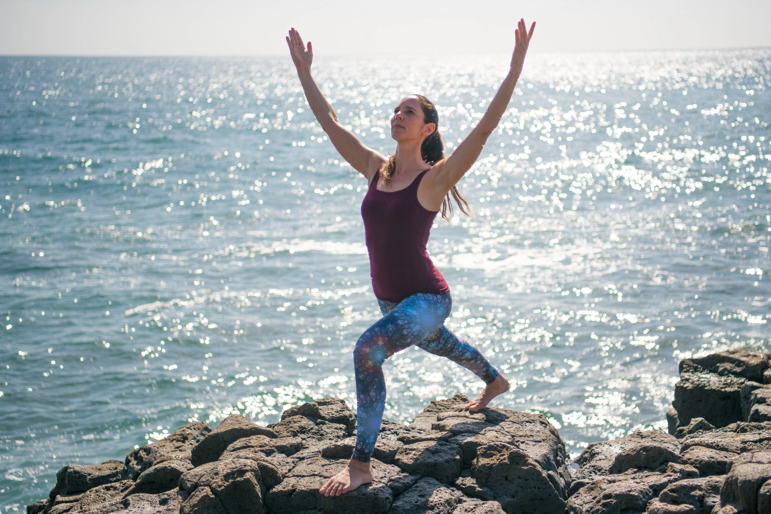 Körperglück - Massagetherapie und Beckenbodentraining - Eine Frau übt die Yoga-Pose „Halbmond-Ausfallschritt“ an einem felsigen Strand am Meer.